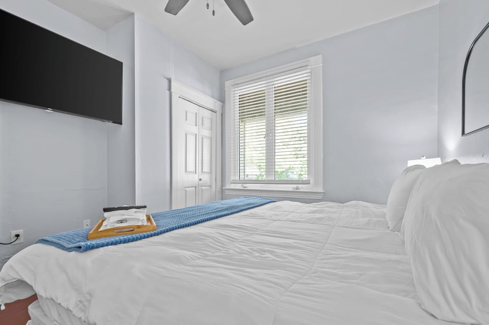 This bedroom includes a white-dressed bed with a blue throw, wall-mounted TV, double-door closet, ceiling fan, broad window with blinds, and arched wall mirror.