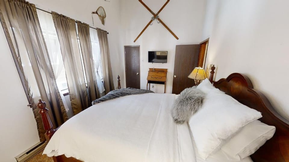 Sink into plush white linens and rustic charm in your cozy bedroom, complete with warm wood accents, a mounted TV, and soft natural light through flowing curtains.