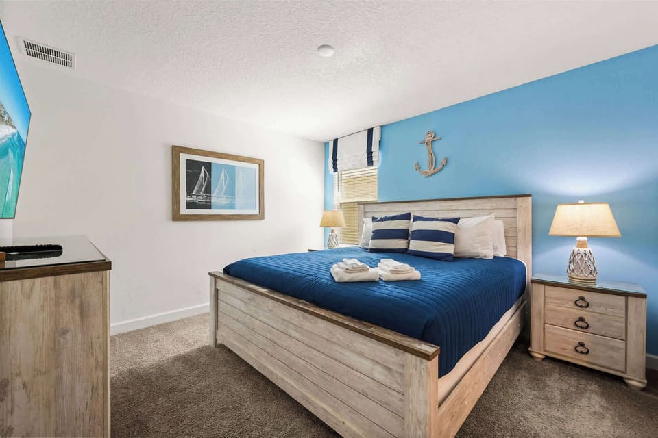 Nautical-themed bedroom with a deep blue quilt, striped pillows, sailboat artwork, anchor decor, and rope-inspired bedside lamps.