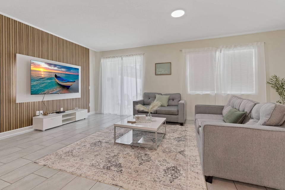 Relax in the bright living room with plush gray sofas and a stunning wooden accent wall that complements the modern decor. Large windows fill the space with natural light, creating a warm and inviting atmosphere for your stay.
