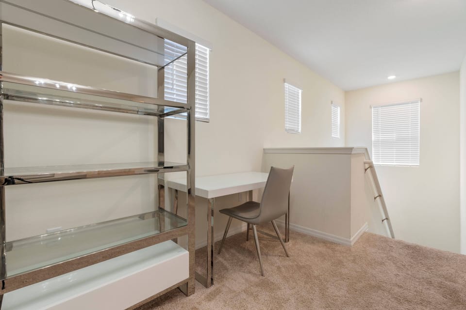 This bright and airy room offers a sleek glass shelving unit and a minimalist desk with a modern chair, ideal for relaxing or working. Large windows let in natural light, creating an inviting atmosphere for your stay.