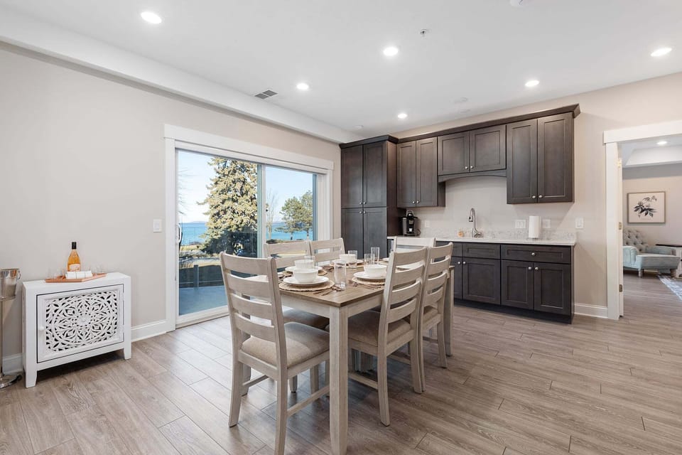 Dining table with white chairs beside dark kitchen cabinets and a bay-view balcony door.