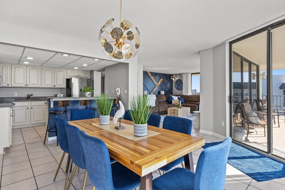 Modern dining area with blue chairs and a wooden table adjacent to a spacious living room and balcony.