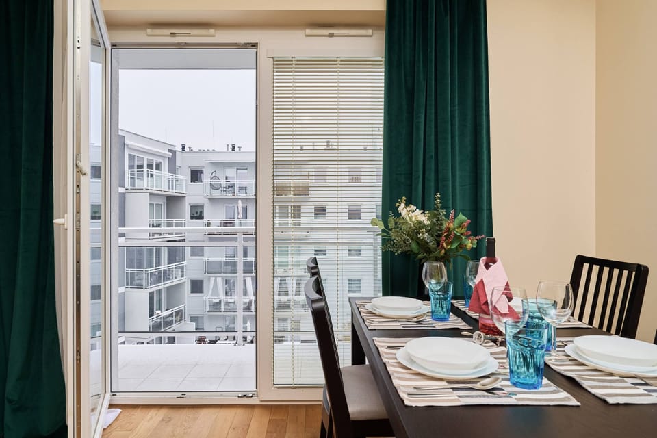 A dining table set for a meal, with colorful plates, wine glasses, and a view of the outside through the balcony doors.