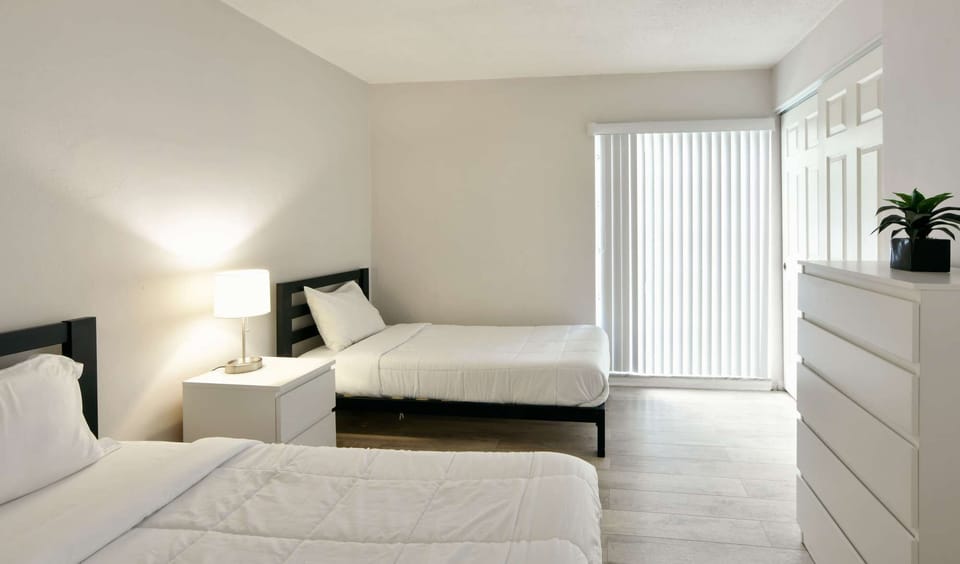 Twin bedroom with dresser, lamp, and large window blinds