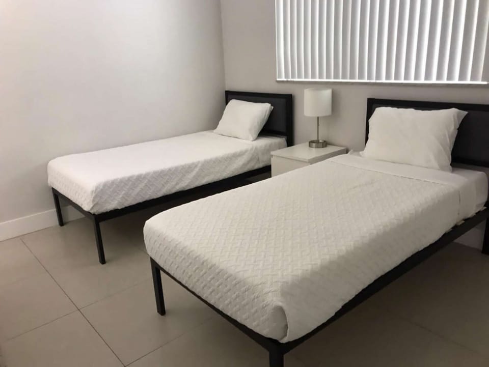 Twin bedroom with two beds, side table, lamp, and window blinds