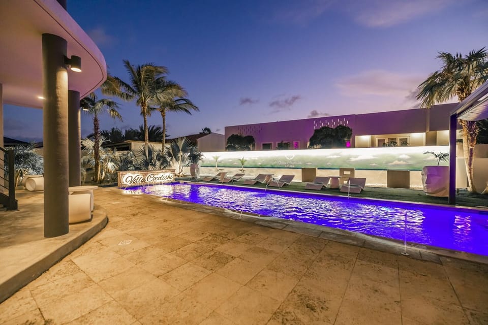 Luxurious villa with a long pool, white lounge chairs with blue towels, palm trees, and modern white architecture creating a bright, tropical retreat.