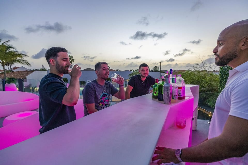 Sunny rooftop lounge with sleek white chairs, a low round table, and cushioned seating framed by glass railings overlooking a pool and palm trees, creating a bright, modern escape.