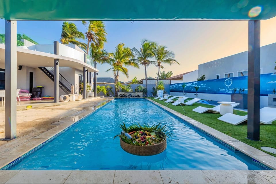Luxurious villa with a long pool, white lounge chairs with blue towels, palm trees, and modern white architecture creating a bright, tropical retreat.