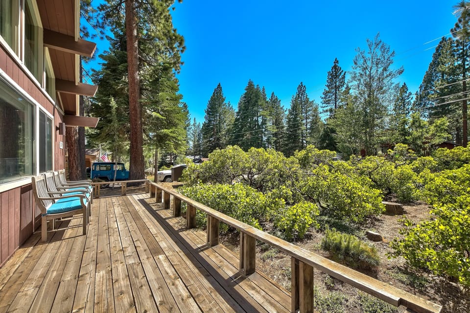 Wooden front deck with railing, lined with cushioned chairs and surrounded by green shrubs and tall pine trees.