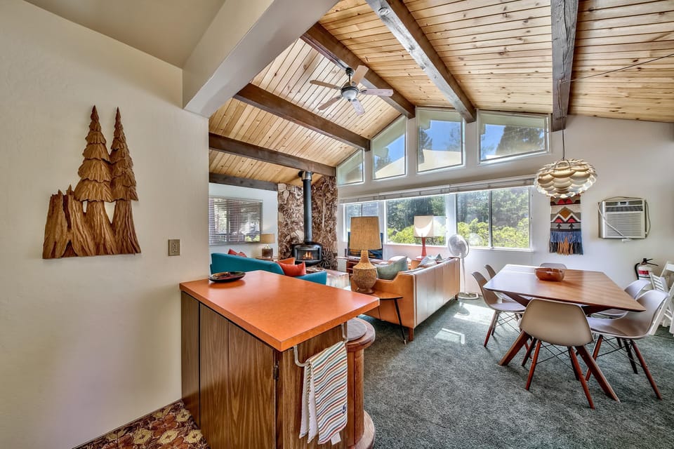 Open living and dining area with vaulted pine ceiling, stone wood stove, orange laminate countertop, and large windows.