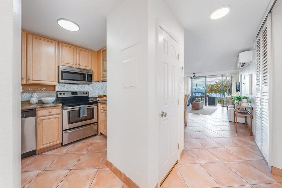 Bright galley‑style kitchen with light wood cabinetry, tile flooring, and a clear view into the open living and dining area.