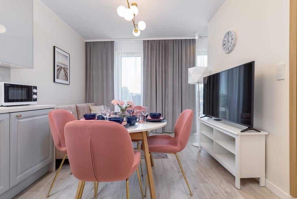 A spacious living room with a large TV, light wooden flooring, and pink accents in the dining area.