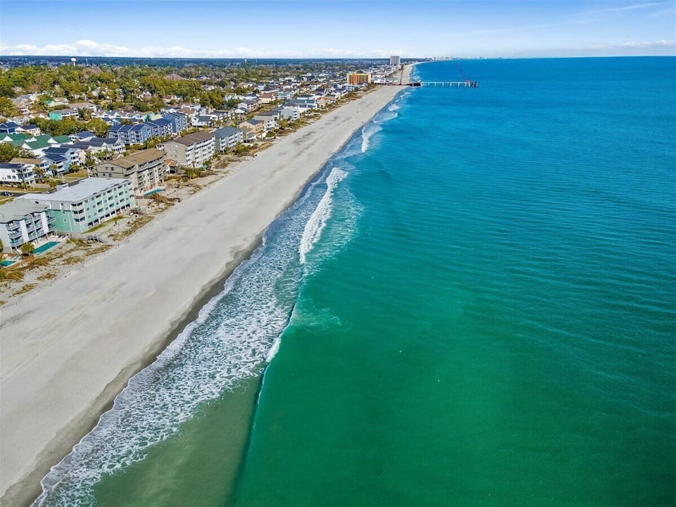 Stunning aerial view of pristine emerald coastline with white sand beach, beachfront properties, and crystal-clear turquoise waters stretching to the horizon.