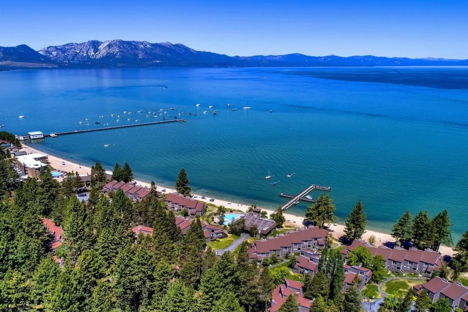 Lakefront resort property nestled along pristine blue waters, with mountain backdrop, private beach access, swimming pool, and multiple vacation units surrounded by pine trees.