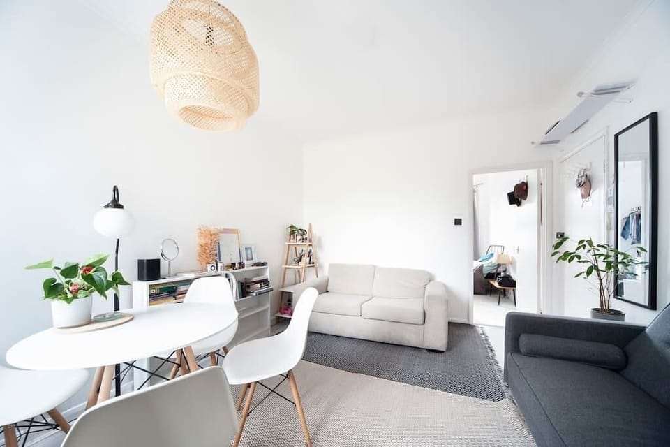 Bright and lively living room featuring two sofas, a dining table with four chairs, a floor lamp, and a mirror. The dining area is positioned near the window. The floor lamp illuminates a cosy corner, perfect for relaxation or reading.