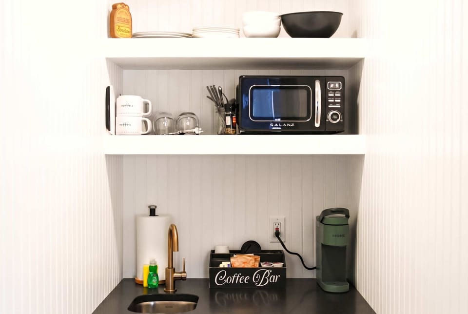 Artist Bluff Kitchenette — Everything You Need, Nothing You Don't. Open shelving with mugs, glasses, and coffee supplies. Microwave, sink, and a countertop that keeps morning simple without taking over the room.