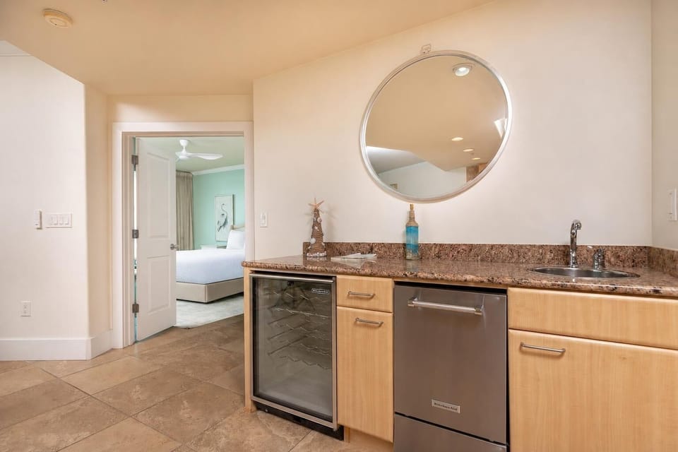 This kitchen features a sleek wet bar with a wine cooler, stylish granite countertops, and a view into the inviting bedroom