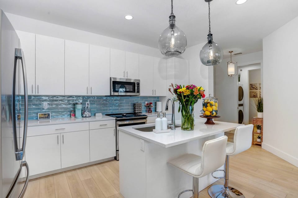 A large open plan kitchen boasts white wood cabinetry, stainless steel appliances, quartz counters, center island and breakfast bar, pendant lighting