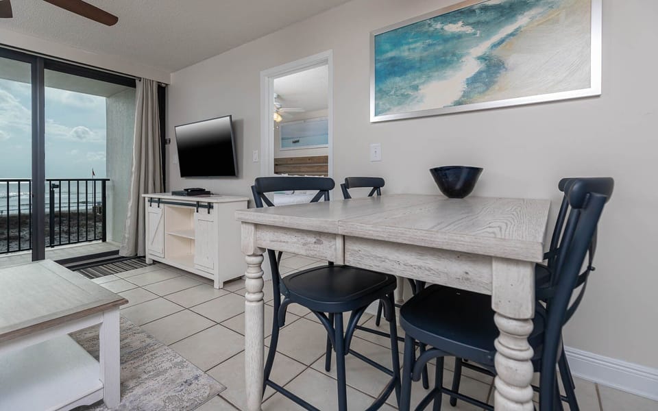 The living area features a large TV, a cozy dining table, and a stunning beachfront view through sliding glass doors