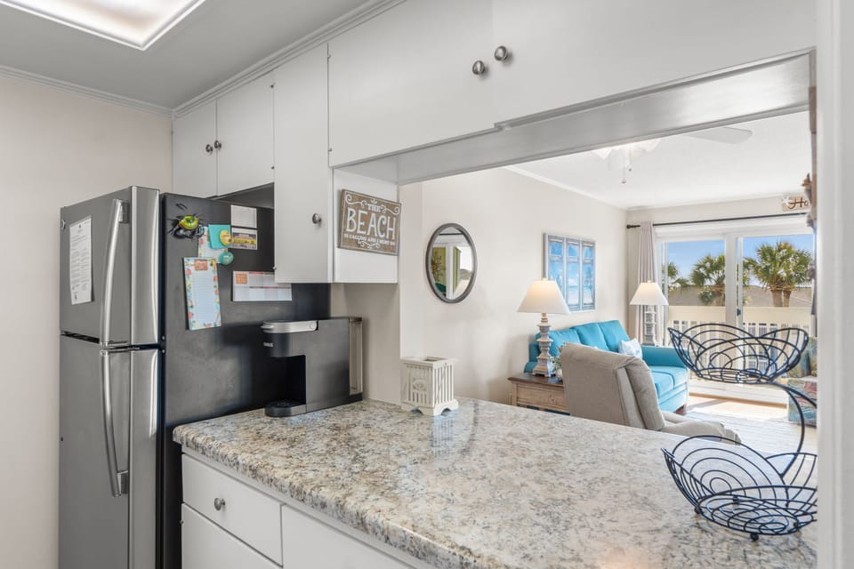Your bright kitchen opens to a coastal living space where palm trees frame ocean views through the windows.