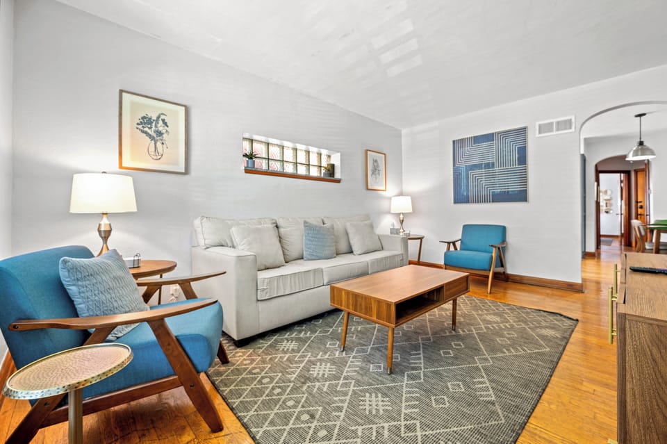 This reverse living-room view shows the sofa, two blue accent chairs, wood coffee table, patterned rug, glass-block window, artwork, and arched hall opening.