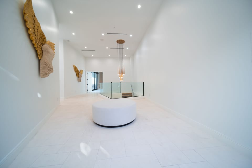 A bright, gallery-like hallway highlights the home's modern design with clean white walls, sculptural décor, and a soft chandelier overhead, creating an elegant transition between living spaces.