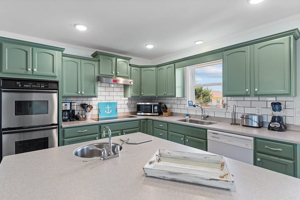 Coastal kitchen with green cabinets, center island sink, double oven, white tile backsplash, dishwasher, window, small appliances, and nautical decor.