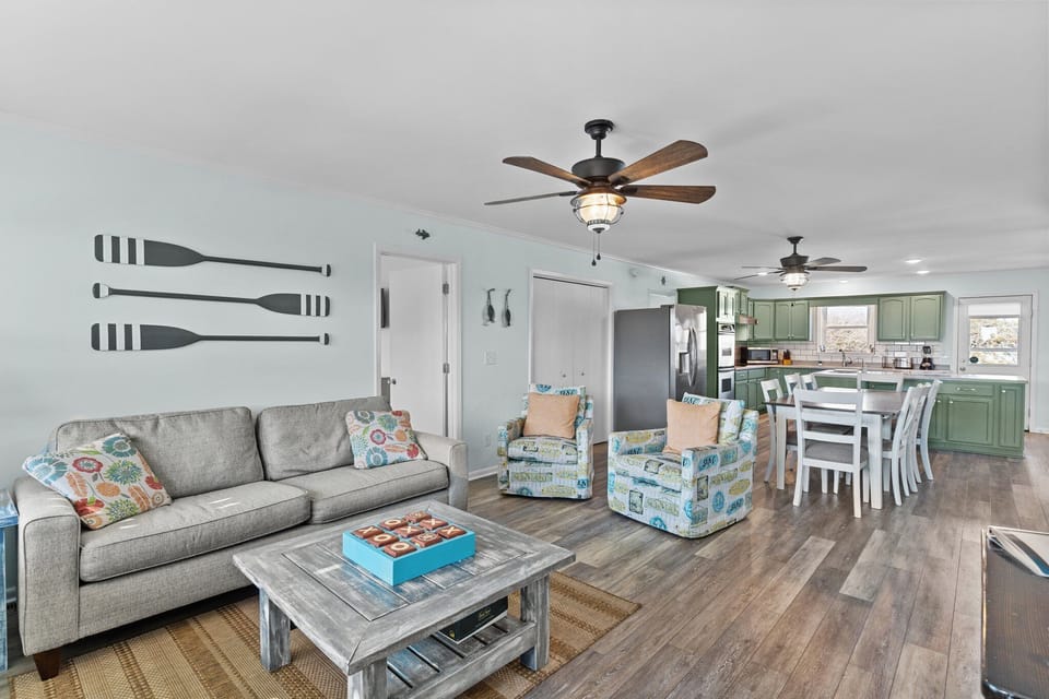 Coastal living room with sofa, patterned chairs, wall TV, ceiling fan, paddle decor, front door, beach house sign, coffee table, and wood floors.