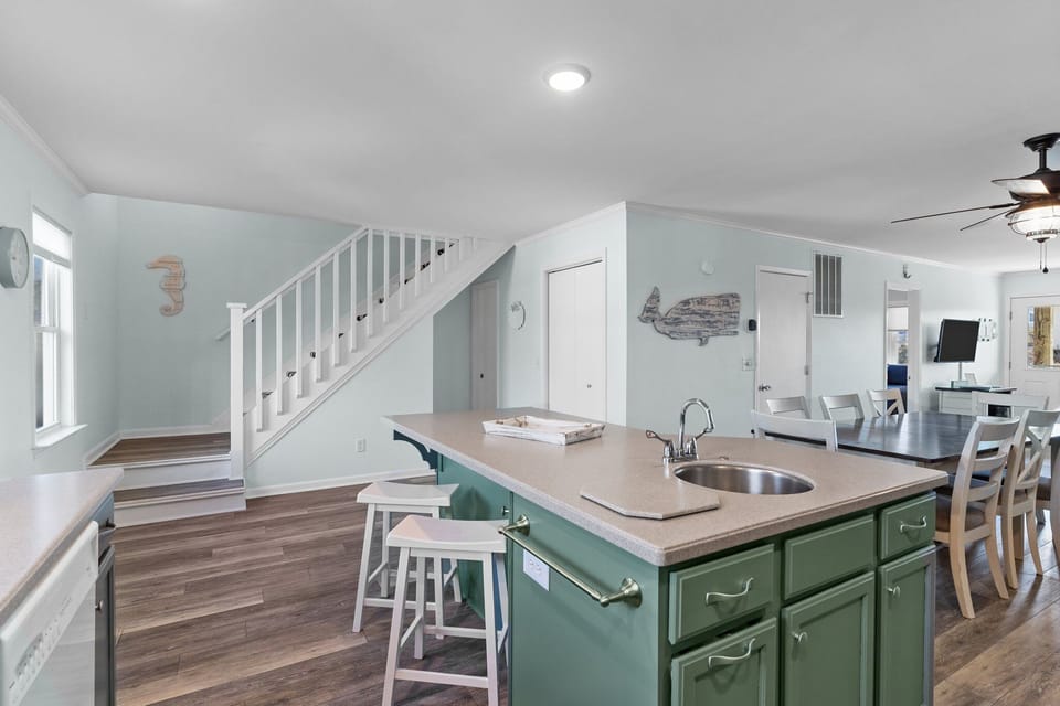 Coastal kitchen with green cabinets, center island sink, double oven, white tile backsplash, dishwasher, window, small appliances, and nautical decor.