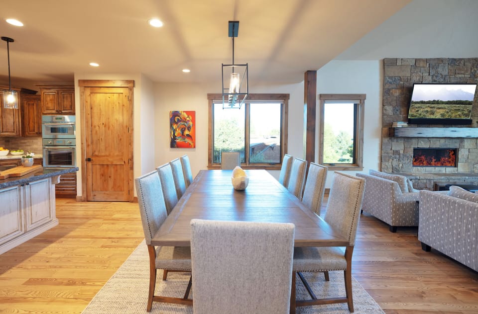 Gather around this elegant dining table where your group can share memorable meals together, surrounded by warm wood tones and natural light streaming through large windows.