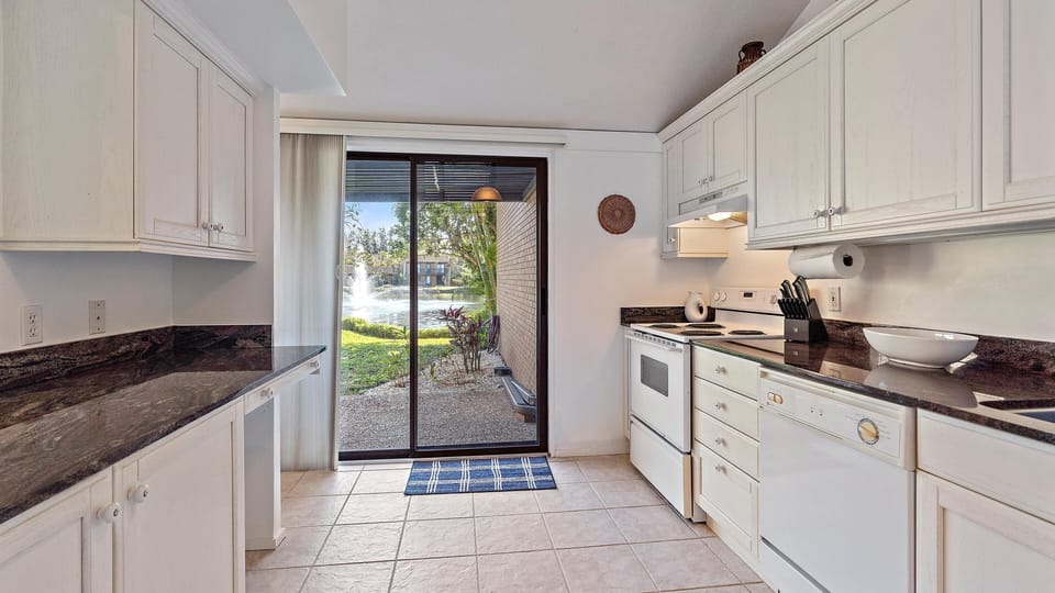 A bright kitchen with white cabinets and black countertops. Sliding glass doors open to a patio with a view of a fountain. The kitchen includes a stove, oven, dishwasher, and microwave.