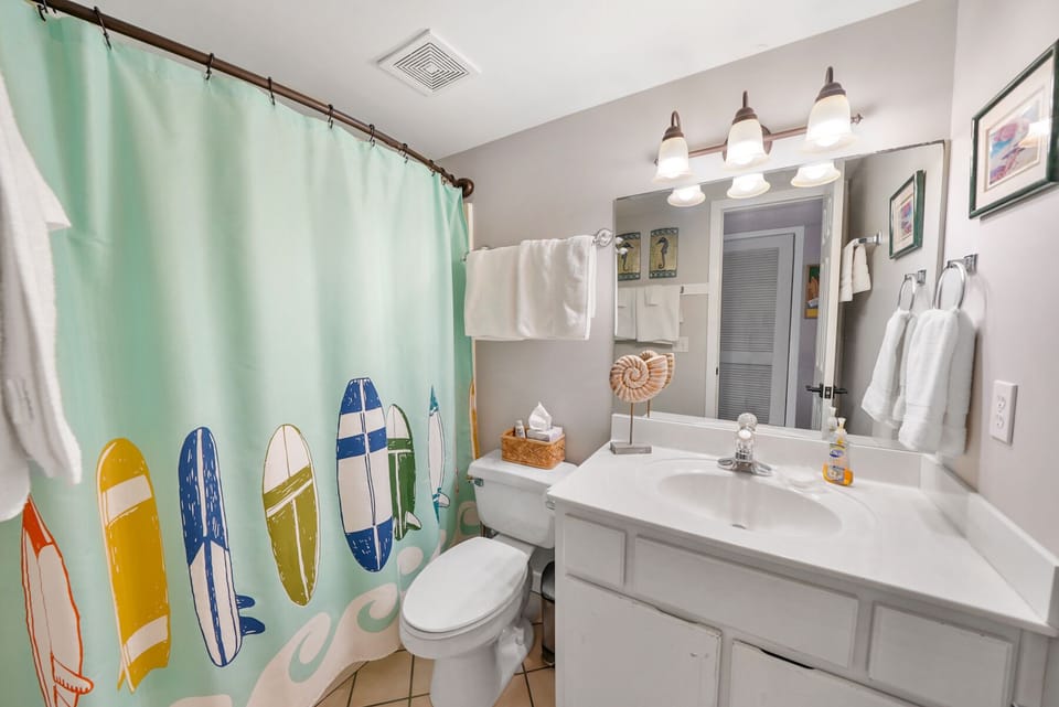 Your beach-themed bathroom features coastal charm with surfboard shower curtains, white vanity, and fluffy towels waiting for your arrival.