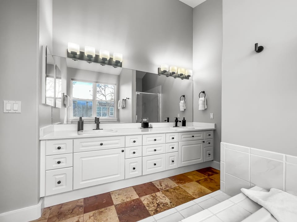 A modern bathroom featuring a double-sink vanity with black fixtures, large mirror, and overhead lighting. There's a window, tiled floor, and a section of the bathtub visible in the foreground.