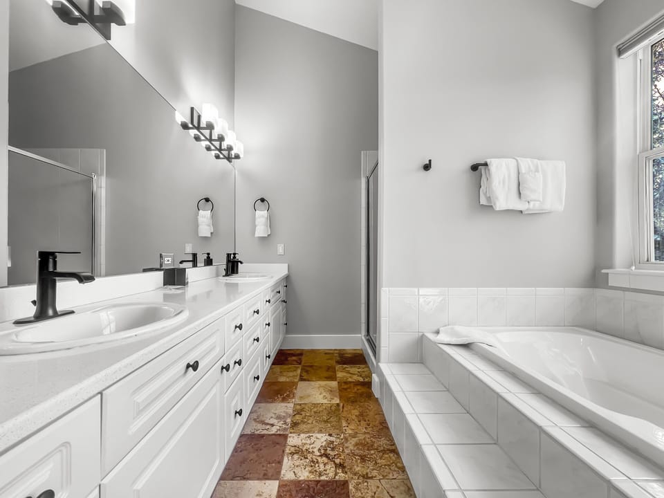 A modern bathroom features a bathtub, a double-sink vanity with black fixtures, a large mirror, white cabinetry, a towel rack with folded towels, and a mixture of tile flooring.