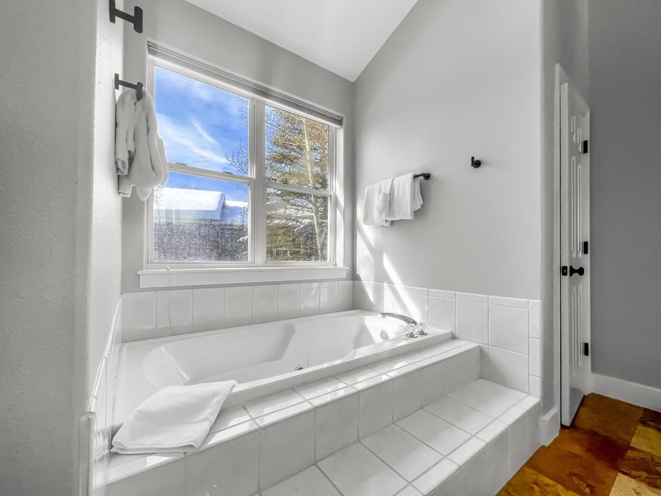 A white tiled bathtub with a towel on the edge sits by a large window with a view of trees and a blue sky. A towel rack with white towels is mounted on the wall beside the window.