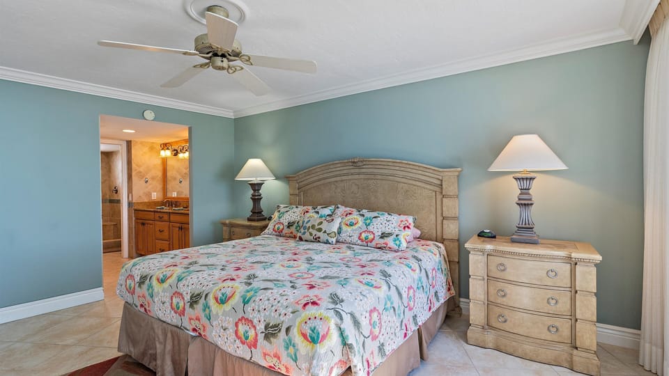 A bedroom with a large bed featuring a colorful floral bedspread, two bedside tables with lamps, a ceiling fan, and a doorway leading to a bathroom. Walls are painted light blue.