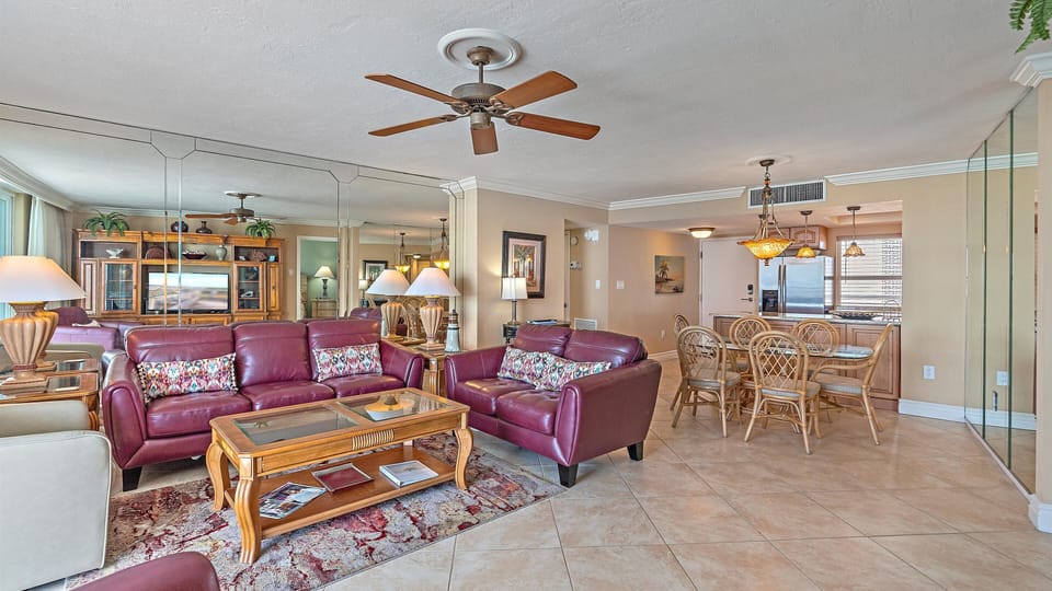 A cozy living room with red leather sofas, glass coffee table, wooden dining set, ceiling fans, and a large mirror wall adjacent to the kitchen. The room is decorated with lamps and framed pictures.