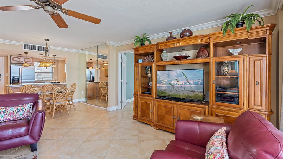 A living room with wooden entertainment center, flatscreen TV, armchairs, tile flooring, ceiling fan, potted plants, and a view into a kitchen with a breakfast bar and wicker chairs.