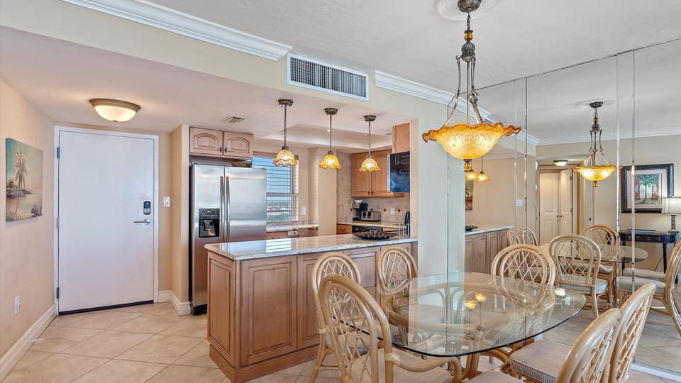 A modern kitchen with a breakfast bar adjacent to a glass dining table with wicker chairs. The space has stainless steel appliances, pendant lights, and a mirrored wall.
