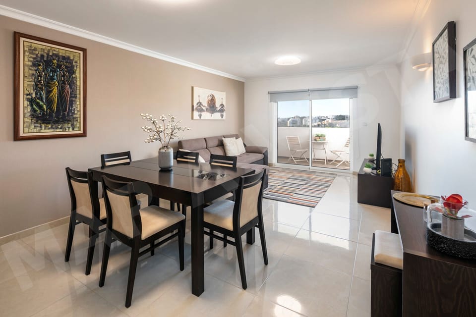 Contemporary dining area in Apartment Mar a Vista with a dark wood table seating six, stylish chairs, vibrant wall art, and natural light flooding in from the balcony, ideal for family meals on your Algarve holiday