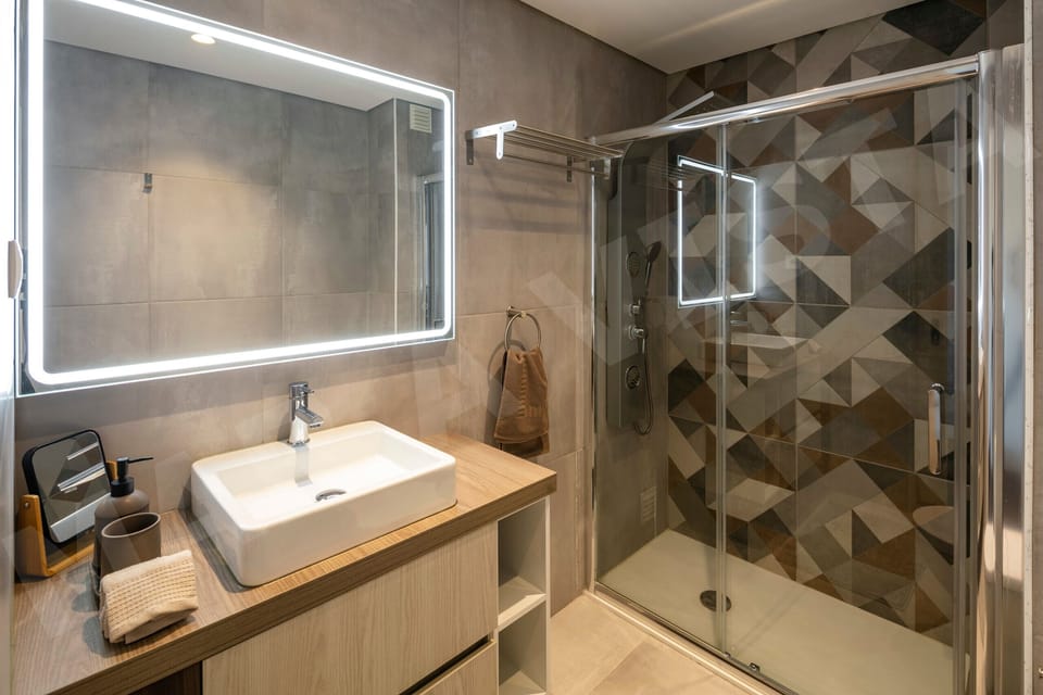 Stylish bathroom in Apartment Mar a Vista featuring an LED-backlit mirror, rectangular basin on a wooden vanity unit, walk-in shower with glass screen, and contemporary geometric patterned dark tiles throughout