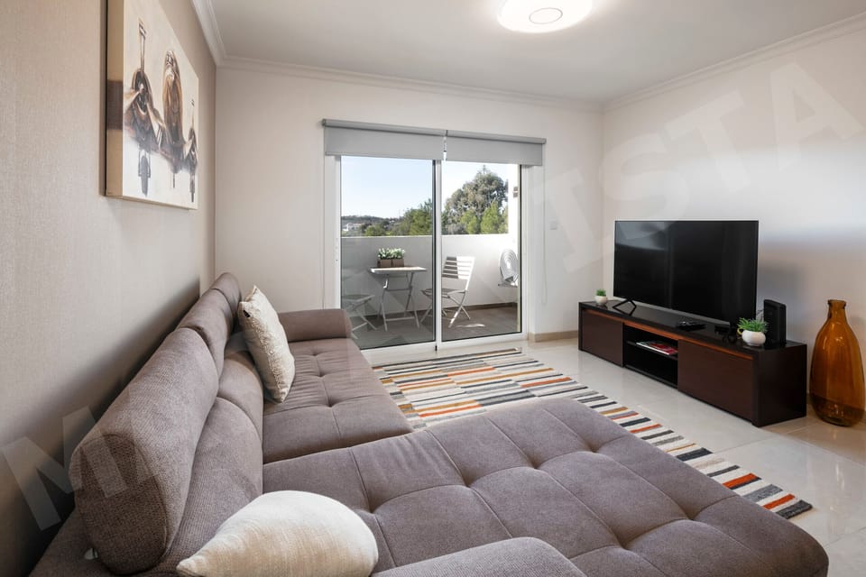 Bright living room in Apartment Mar a Vista with a large grey corner sofa, flat-screen TV, modern wall art, and sliding doors opening onto a furnished balcony with views across the Algarve landscape