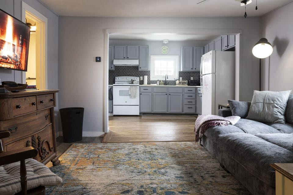 Farmhouse Living Room – The farmhouse living room features a comfortable couch, TV, and curated artwork, along with a side table for added convenience. The open layout provides easy access to the kitchen and bedrooms, creating a welcoming and functional g