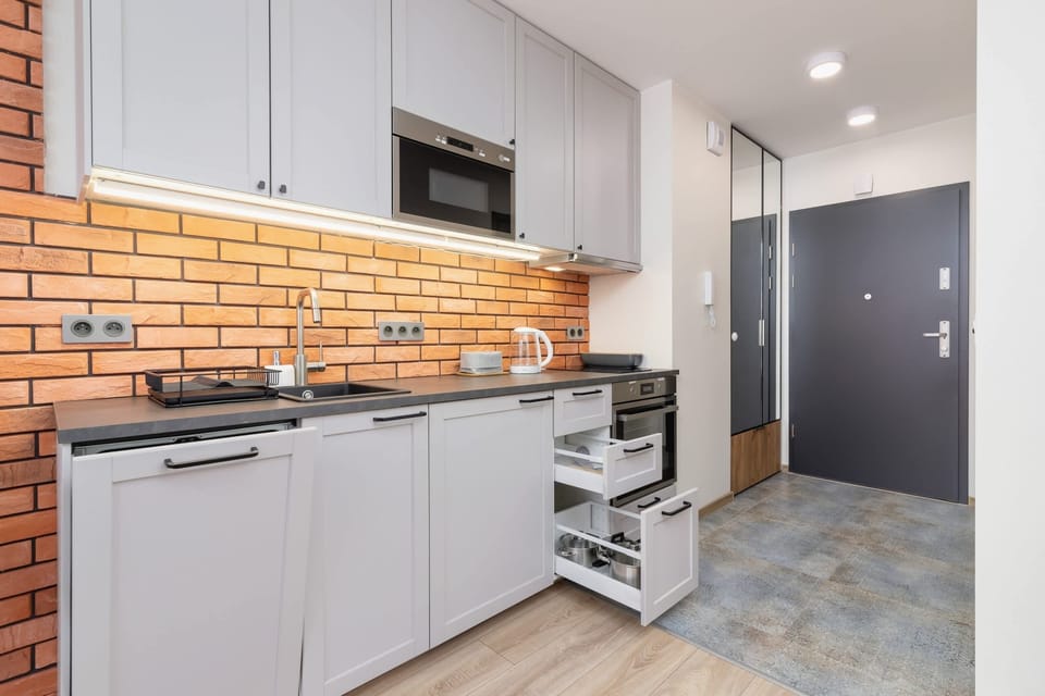A kitchenette with gray cabinets, a brick backsplash, and modern appliances, leading to the entrance.
