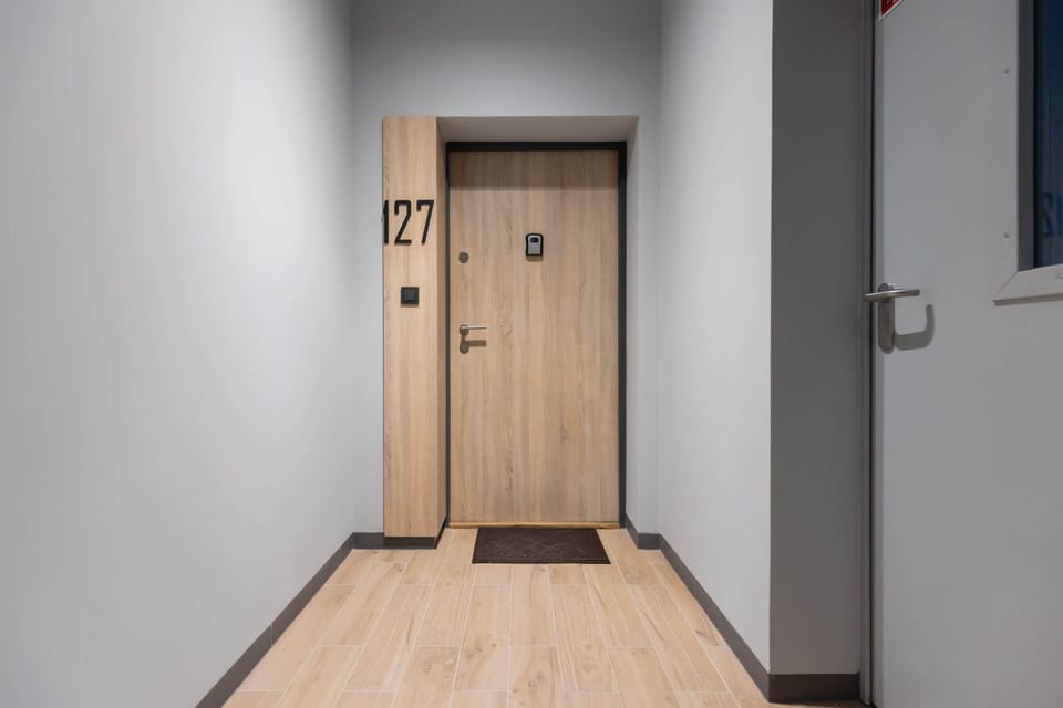 A hallway leading to the apartment entrance, featuring a wooden door and clean, modern finishes.

