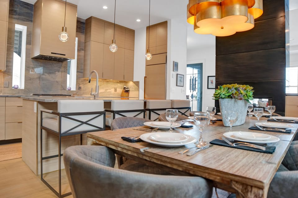 The dining area overlooking a kitchen with light colored cabinets, fridge with light wood matching door and grey backsplash.