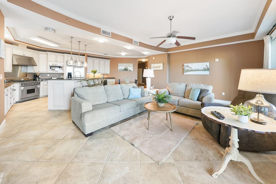 Relax in your spacious open-concept living space with a stylish sectional sofa, modern kitchen with white cabinets, and coastal artwork adorning warm beige walls.
