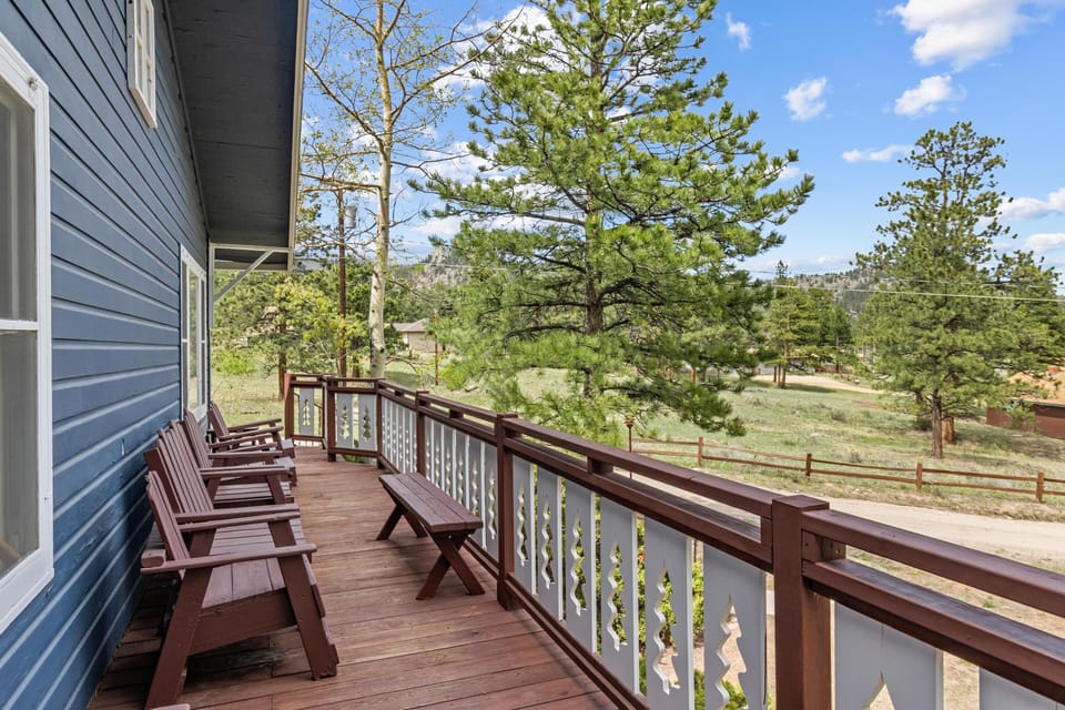 Wooden deck with brown Adirondack chairs and bench overlooks pine trees and open meadow under blue sky.