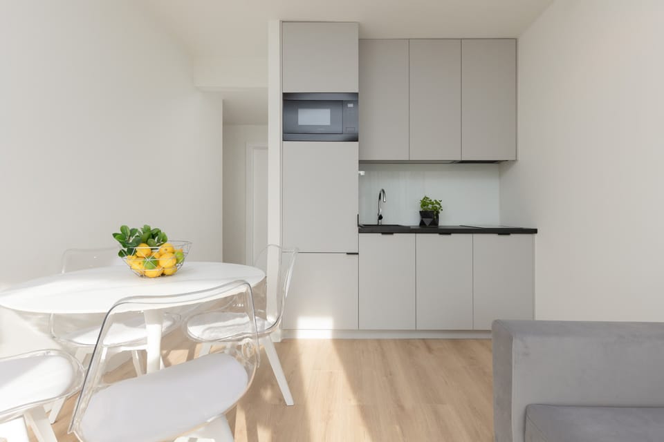 A minimalist dining corner with white furniture and a small kitchen.
